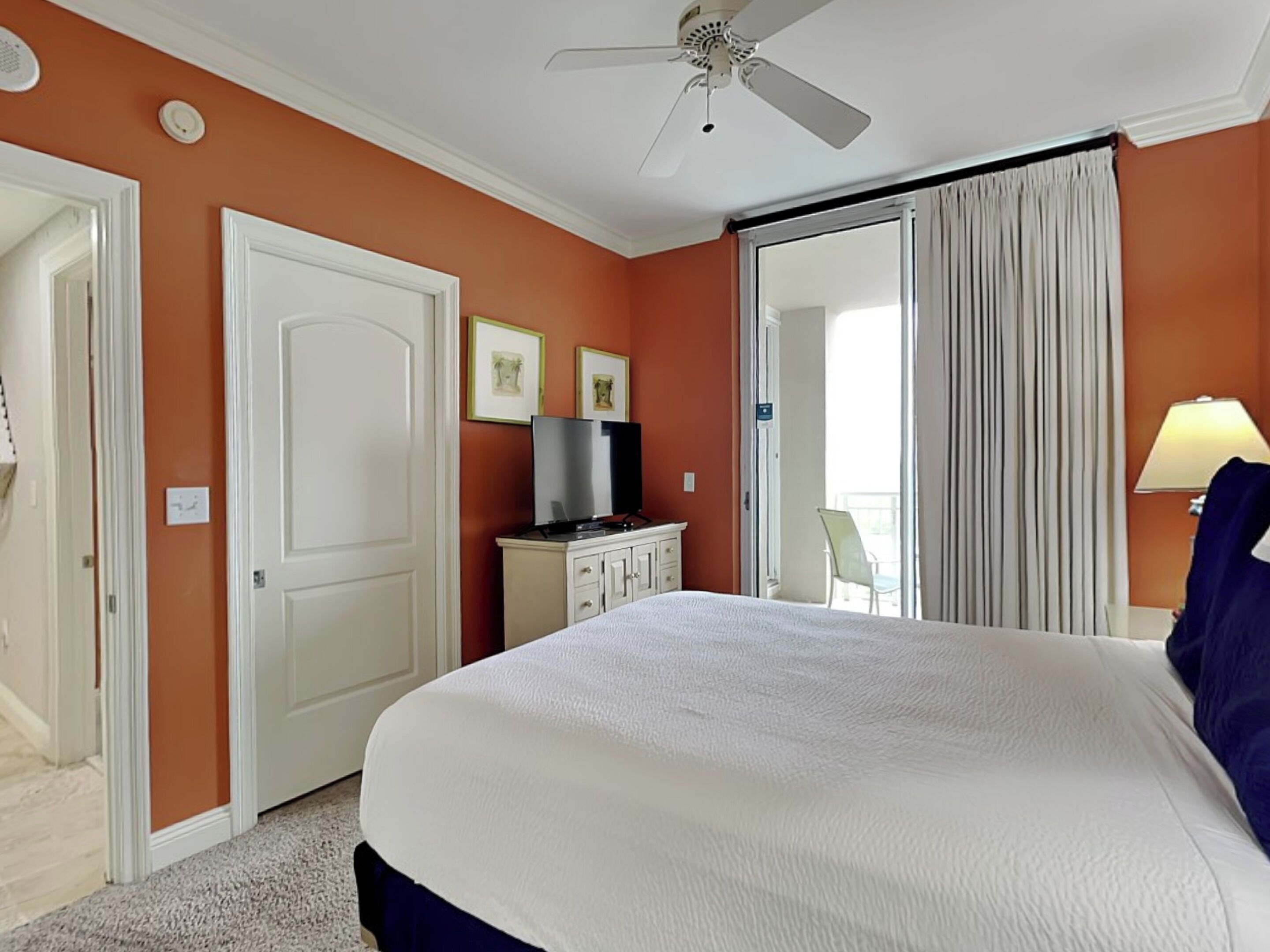 A hotel room with a large bed, a ceiling fan, a television on a cabinet, and a view of the outside through a sliding door.