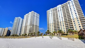 A beachfront hotel with multiple high-rise buildings, a sandy beach, and palm trees. - Indigo East #705 | Luxe Gulf Front Condo + Amenities (Perdido Key)