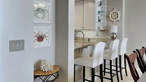 A modern kitchen with white cabinets, a marble countertop, and a bar area with white chairs. - Indigo East #705 | Luxe Gulf Front Condo + Amenities (Perdido Key)