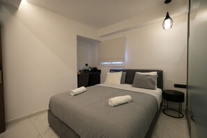 A modern bedroom with a large bed, bedside tables, a desk, and a window with blinds. - Signature Suites - Stadiou (Athens)