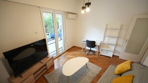 Ein modernes Wohnzimmer mit Flachbildfernseher, einem kleinen Tisch, einer Couch mit Kissen, einem Schreibtisch und einem Sessel.