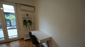 Ein Zimmer mit weißem Schreibtisch, schwarzem Bürostuhl, Holzboden im Fischgrätmuster, einer weißen Regaleinheit mit Pflanzen und einer Glastür, die zu einem Außenbereich führt.