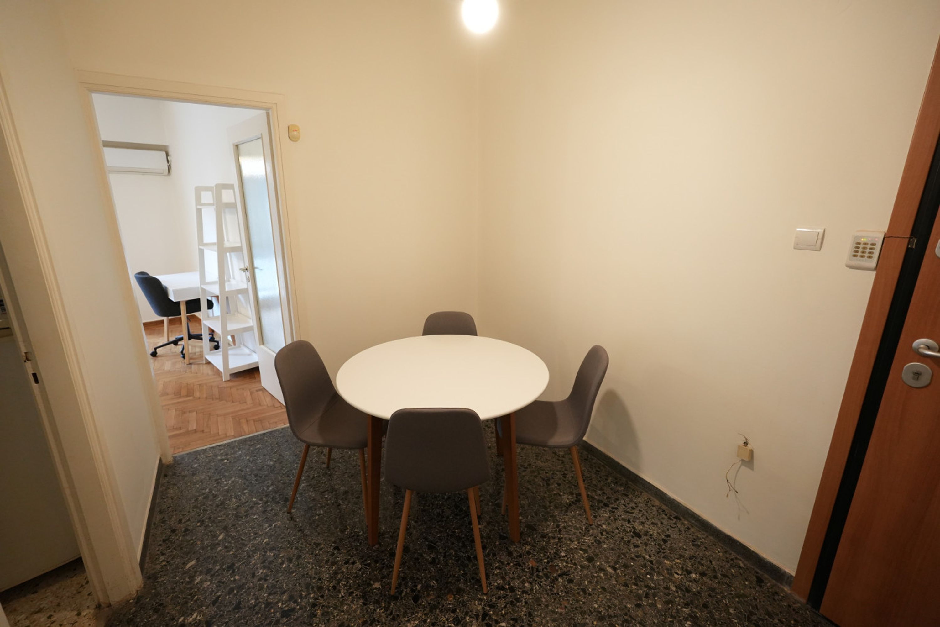A small, private meeting room with a round table and four chairs, carpeted floor, and a door leading to a hallway with a white chair and a desk.