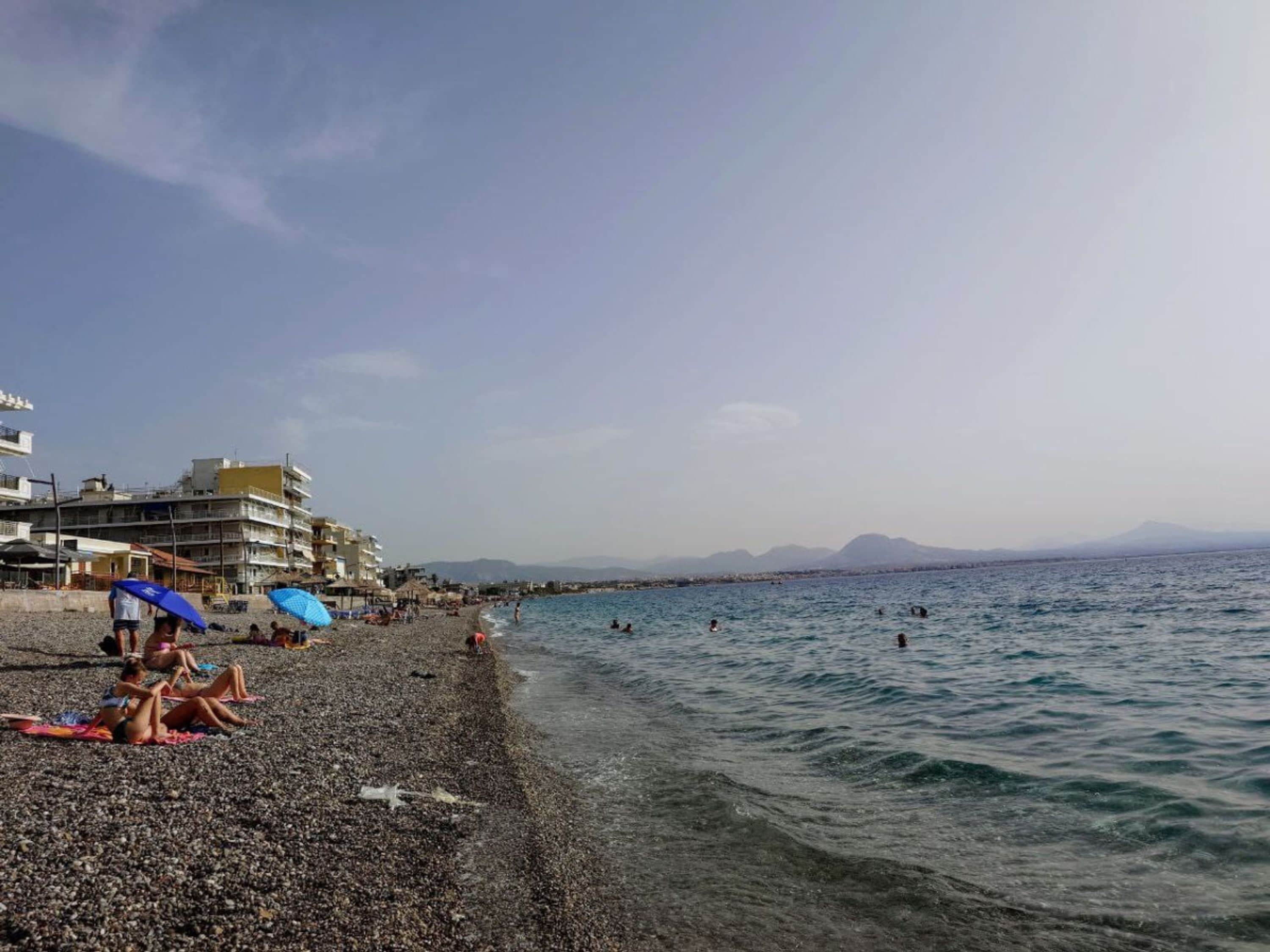 Une plage de galets où des gens se prélassent au soleil et se baignent, avec des bâtiments en arrière-plan et des montagnes à l’horizon.