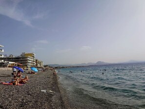 A pebbled beach with people sunbathing and swimming, buildings in the background, and mountains in the distance. - Couples Cozy Seaside Studio (Loutraki-Agioi Theodoroi)