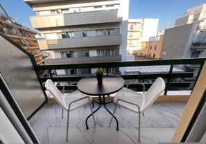 A balcony with a round table and two white chairs, overlooking a cityscape with modern buildings. - Couples Cozy Seaside Studio (Loutraki-Agioi Theodoroi)