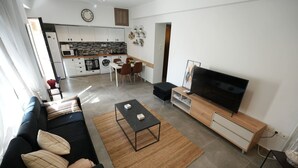A modern living room with a black sofa, a coffee table, and a TV on a wooden cabinet. There is a kitchen in the background with white cabinets and a dining area with a table and chairs. - Lovely Front Garden 2 Bdrm Nea Smyrni (Nea Smirni)