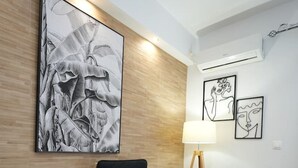 A modern room with a wooden accent wall, a black upholstered chair with a striped pillow, a floor lamp, and a small table with a lamp. - Lovely Front Garden 2 Bdrm Nea Smyrni (Nea Smirni)