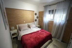 Ein Schlafzimmer mit einem hölzernen Kopfende, einem Bett mit roter Decke, einem Nachttisch und einem Fenster mit Vorhängen.
