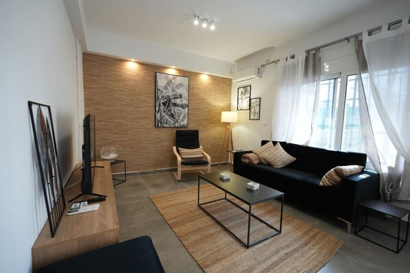 A modern living room with a black sofa, a wooden accent wall, and a large window with sheer curtains. - Lovely Front Garden 2 Bdrm Nea Smyrni (Nea Smirni)