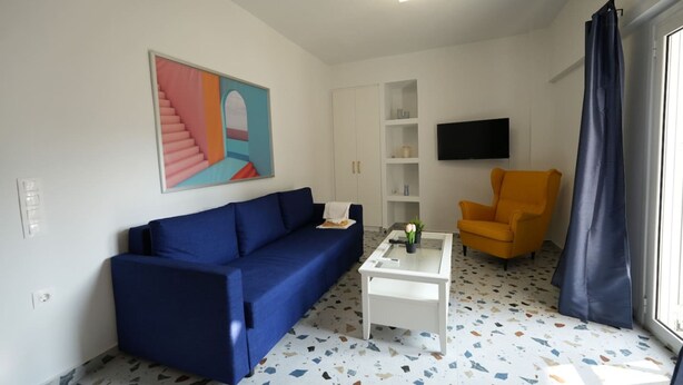 Una sala de estar con un sofá azul, una mesa de centro blanca, un sillón amarillo, un televisor montado en la pared y un piso con diseño.