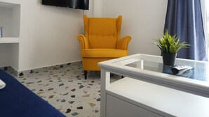 Una habitación con un televisor blanco montado en la pared, un sillón amarillo, un escritorio blanco con una planta y un sofá azul.