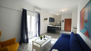 Una sala moderna con un sofá azul, un sillón amarillo y una mesa de centro blanca. En el fondo, se observa una cocina con un taburete de bar y un refrigerador.