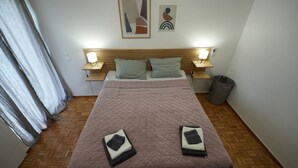A hotel room with a bed, two pillows, a bedside table with a lamp, a trash bin, and a wall with abstract art. - Loutraki Seaview Firstline (Loutraki-Agioi Theodoroi)