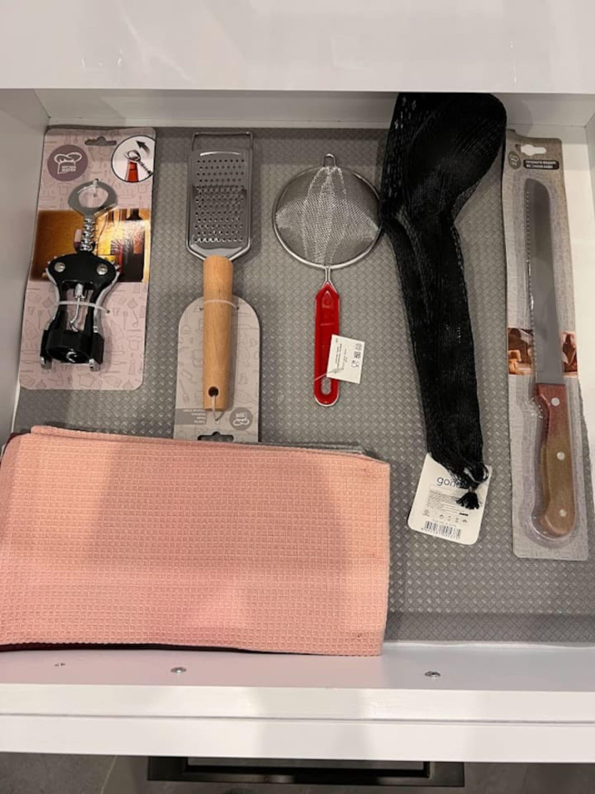 A drawer containing kitchen utensils including a grater, strainer, and a black hair tie.