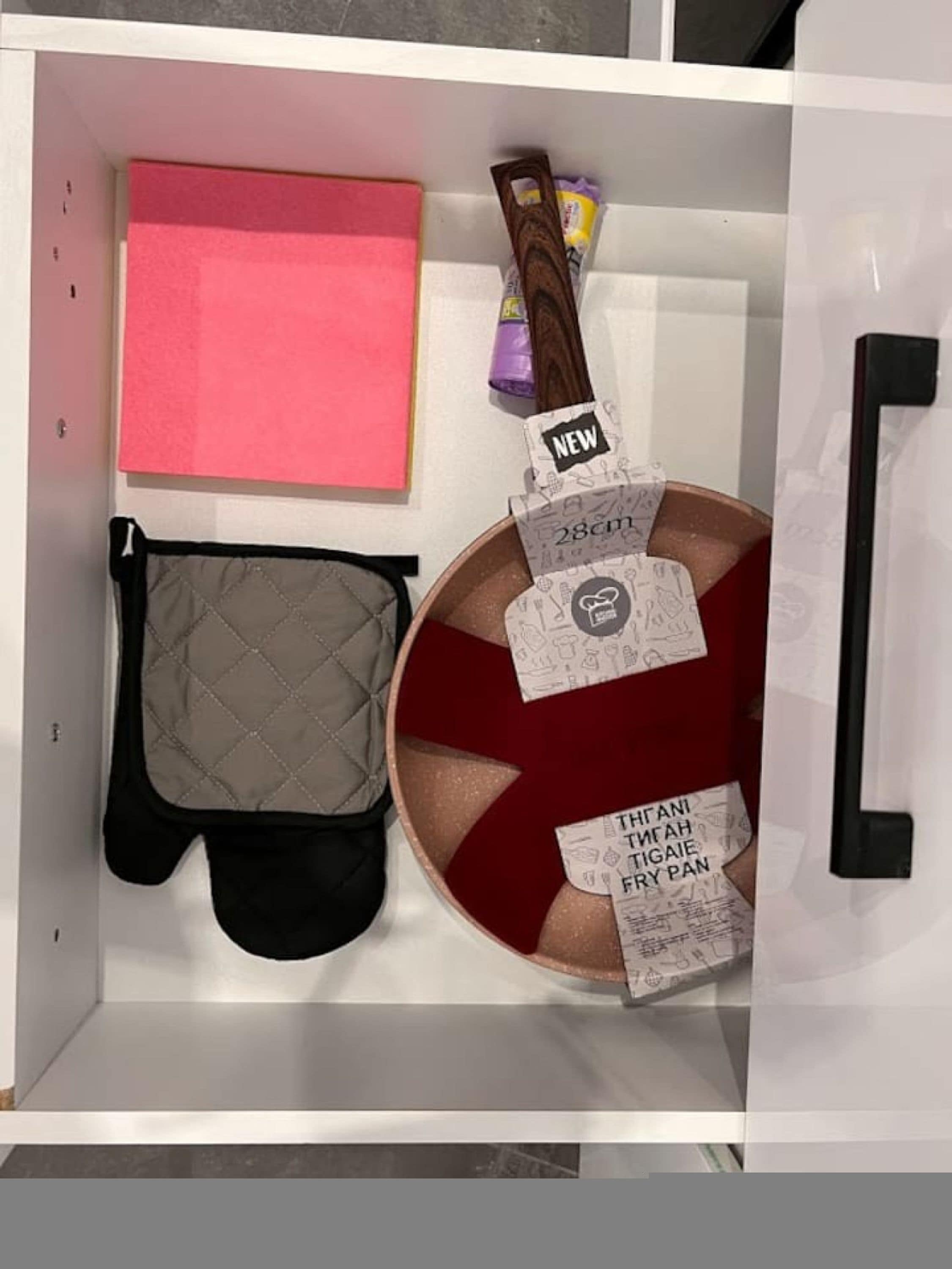 A kitchen drawer with a pink cutting board, a black oven mitt, a red pot, and a new spice rack.