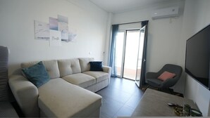 A modern living room with a sofa, a coffee table, and a TV. There is a sliding glass door leading to an outdoor area. - Loutraki Seaview Firstline (Loutraki-Agioi Theodoroi)