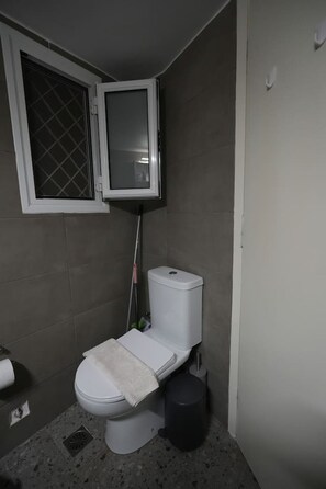 A small bathroom with a toilet, a bidet, a trash bin, and a window with a metal grid. - Best View Loutraki 2 Bedroom (Loutraki-Agioi Theodoroi)
