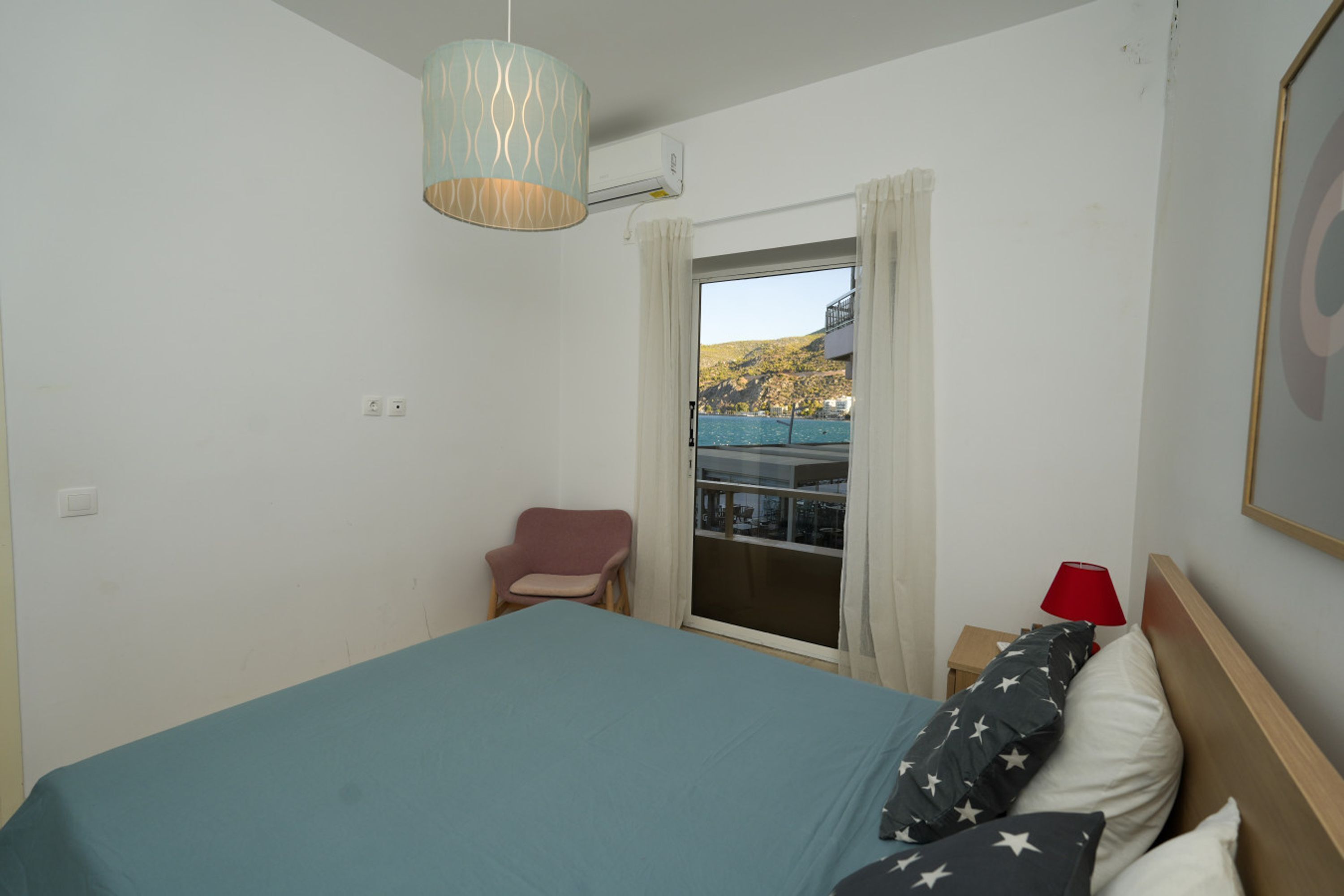 A hotel room with a bed, a chair, a red lamp, and a view of a balcony with a railing.