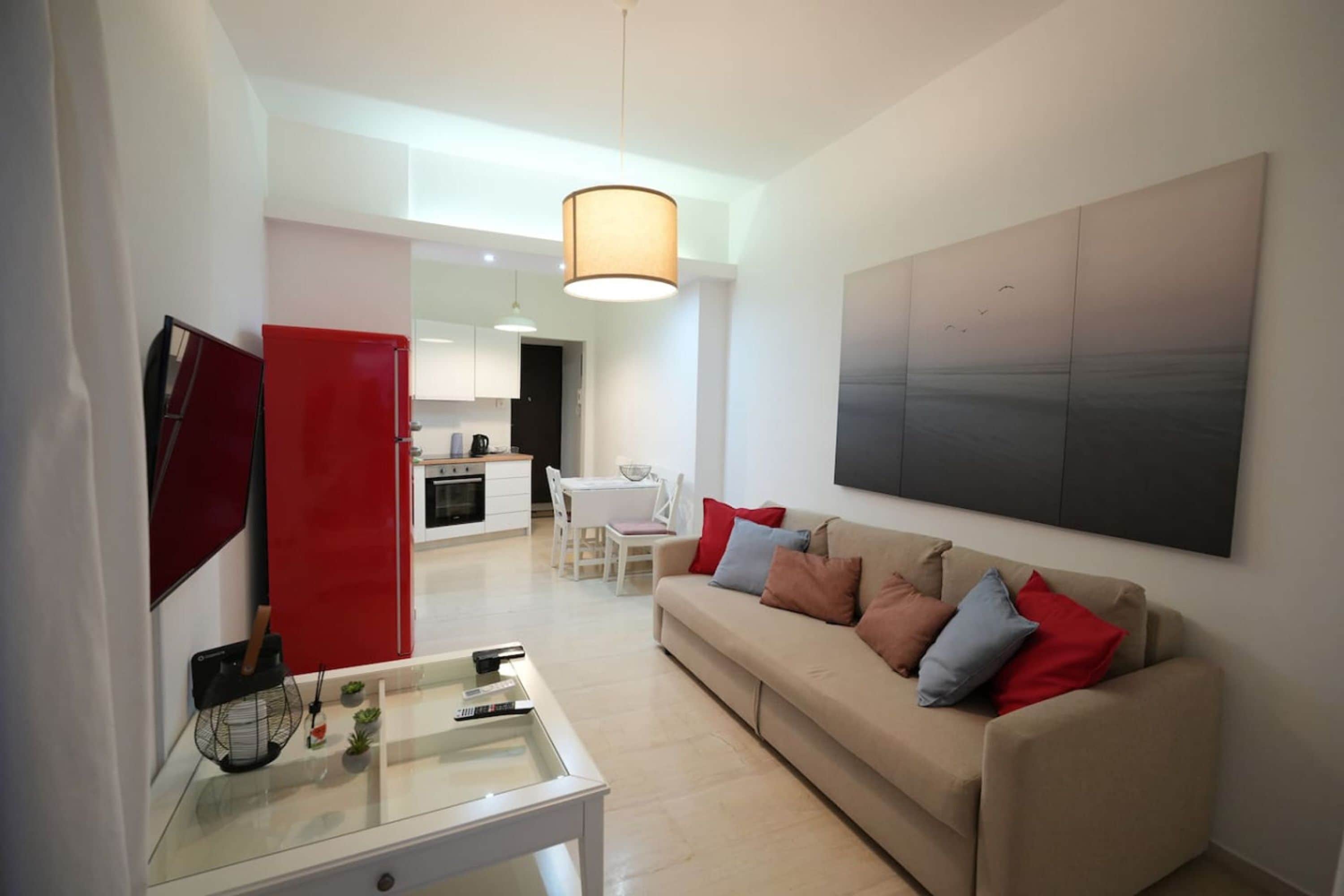 Un salon moderne avec un canapé beige, une table basse en verre et un meuble rouge. On aperçoit au fond une cuisine avec des meubles blancs et un coin repas.