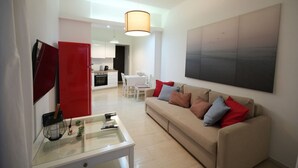 A modern living room with a beige sofa, a glass coffee table, and a red cabinet. There is a kitchen in the background with white cabinets and a dining area. - Best View Loutraki 2 Bedroom (Loutraki-Agioi Theodoroi)