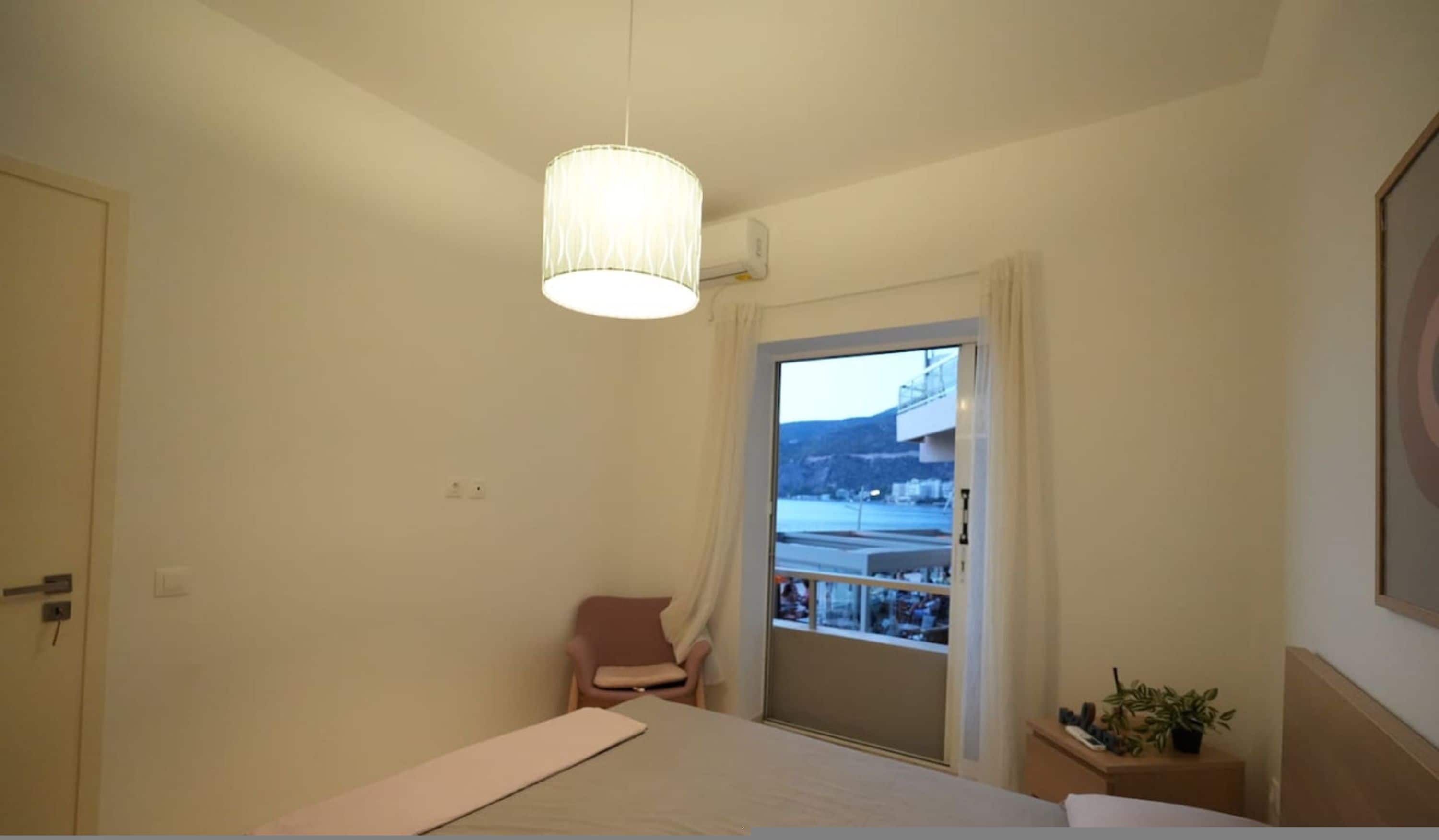 Un dormitorio con cama, una silla, una mesita con una planta y una ventana que ofrece vistas a un balcón y edificios.