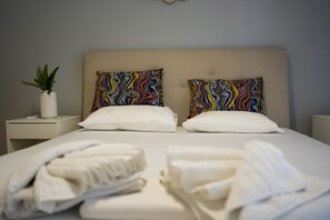 A neatly made bed with two patterned pillows, a bedside plant, and a stack of towels on a white nightstand. - Ilysia s Retreat Cozy 1 Bdrm Apartment (Athens)