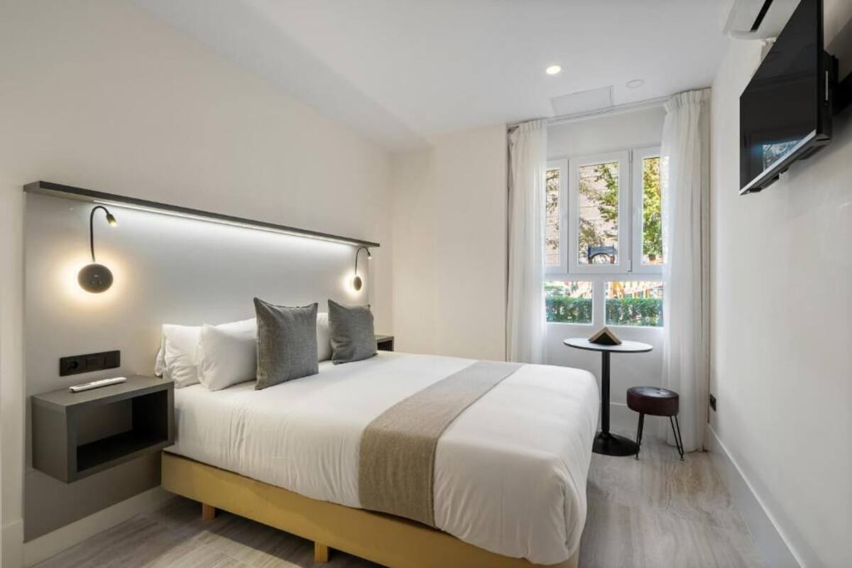 Ein modernes Hotelzimmer mit einem großen Bett, Nachttischen, einem an der Wand montierten Fernseher und einem Fenster mit Blick auf Grünflächen.