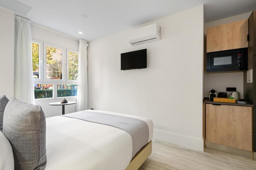 Habitación de hotel con cama, televisor montado en la pared, una kitchenette pequeña con microondas y una ventana con cortinas.