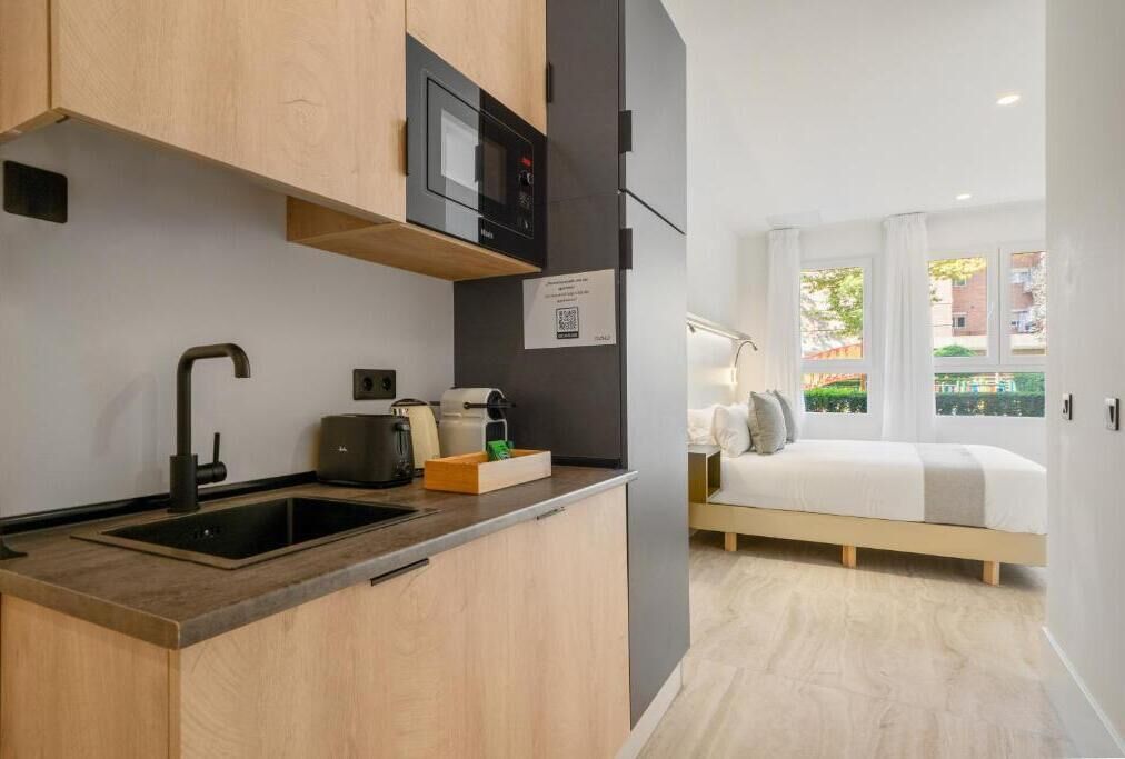 Cocina compacta con microondas, lavabo y cafetera. Se ve una cama con vistas al exterior.
