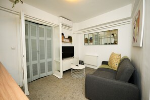 A living room with a grey sofa, a television on a white cabinet, a round coffee table, and a door leading to a closet. - The Extravagant Studio in Pagkrati Near Stadium (Athens)