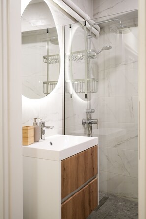 A modern bathroom with a white vanity, wooden cabinet, and a glass-enclosed shower area. - The Extravagant Studio in Pagkrati Near Stadium (Athens)