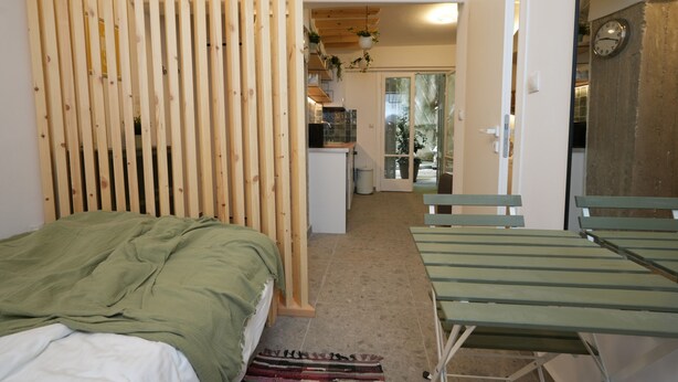 A bedroom with a bed, a wooden partition, a green sofa, and a view of a kitchen and balcony. - The Extravagant Studio in Pagkrati Near Stadium (Athens)