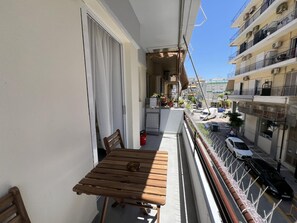 A balcony with a wooden table and chairs, overlooking a street with parked cars and buildings. - Loutraki Haven 1 Bdrm 3 min to Beach (Loutraki-Agioi Theodoroi)
