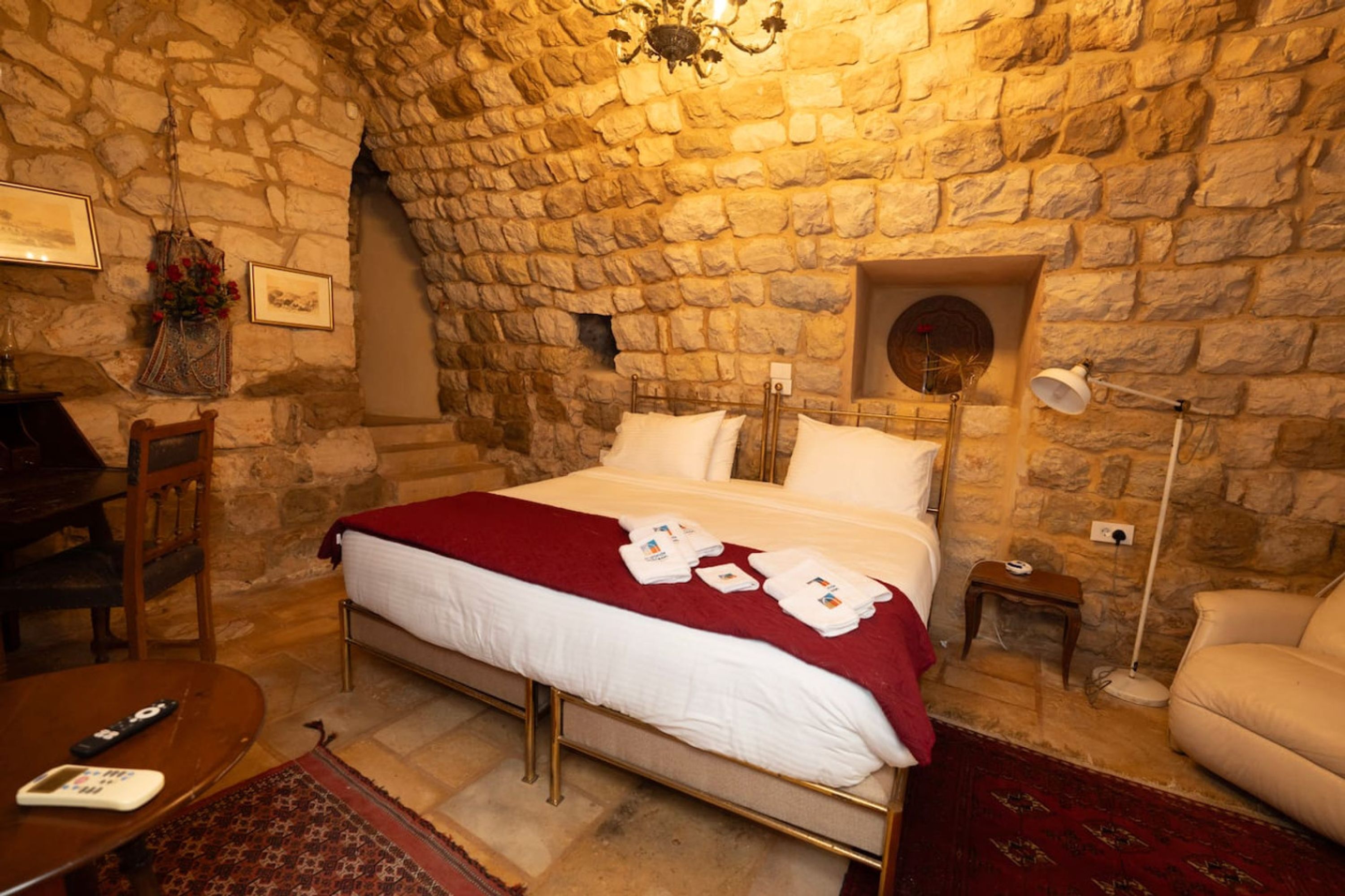 A room with a stone wall, a bed with white bedding, a red throw, a small bedside table with a lamp, and a chair.