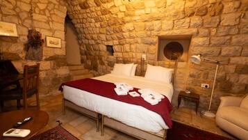 A room with a stone wall, a bed with white bedding, a red throw, a small bedside table with a lamp, and a chair.