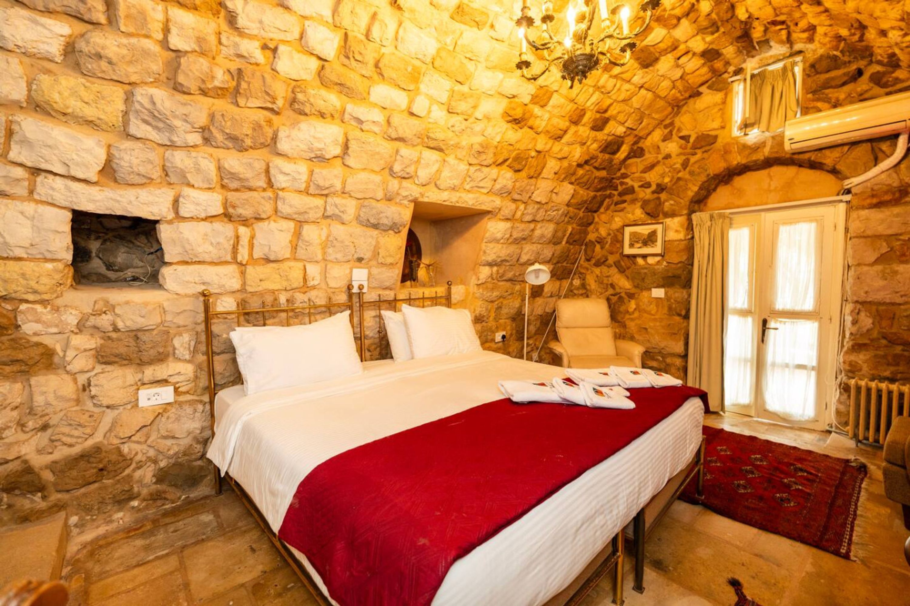 A bedroom with a stone wall, a bed with white bedding, a red throw blanket, and a wooden chair.
