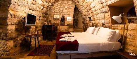 A stone-walled bedroom with a bed, a desk, and a television.