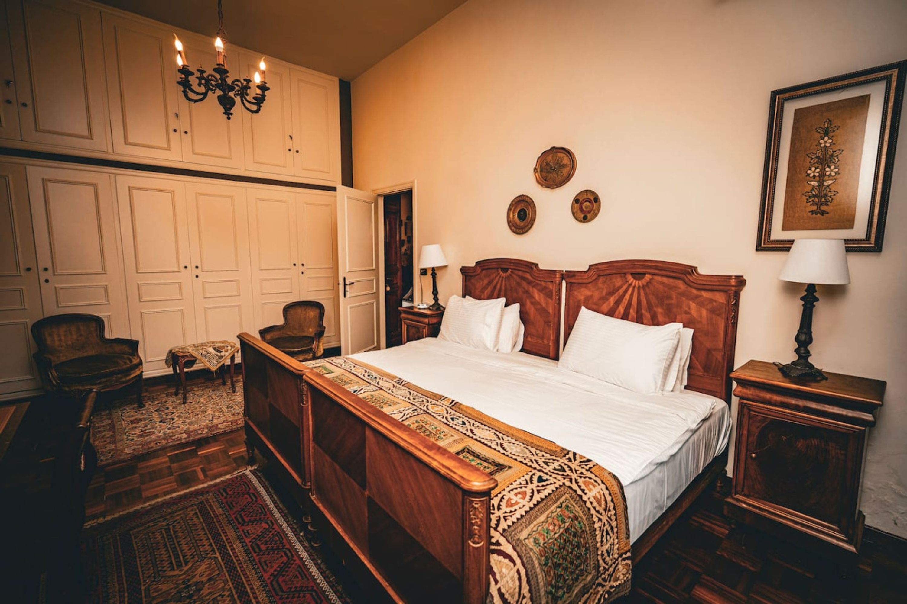 A bedroom with a large wooden bed, two bedside tables, a chair, a wardrobe, and a chandelier.
