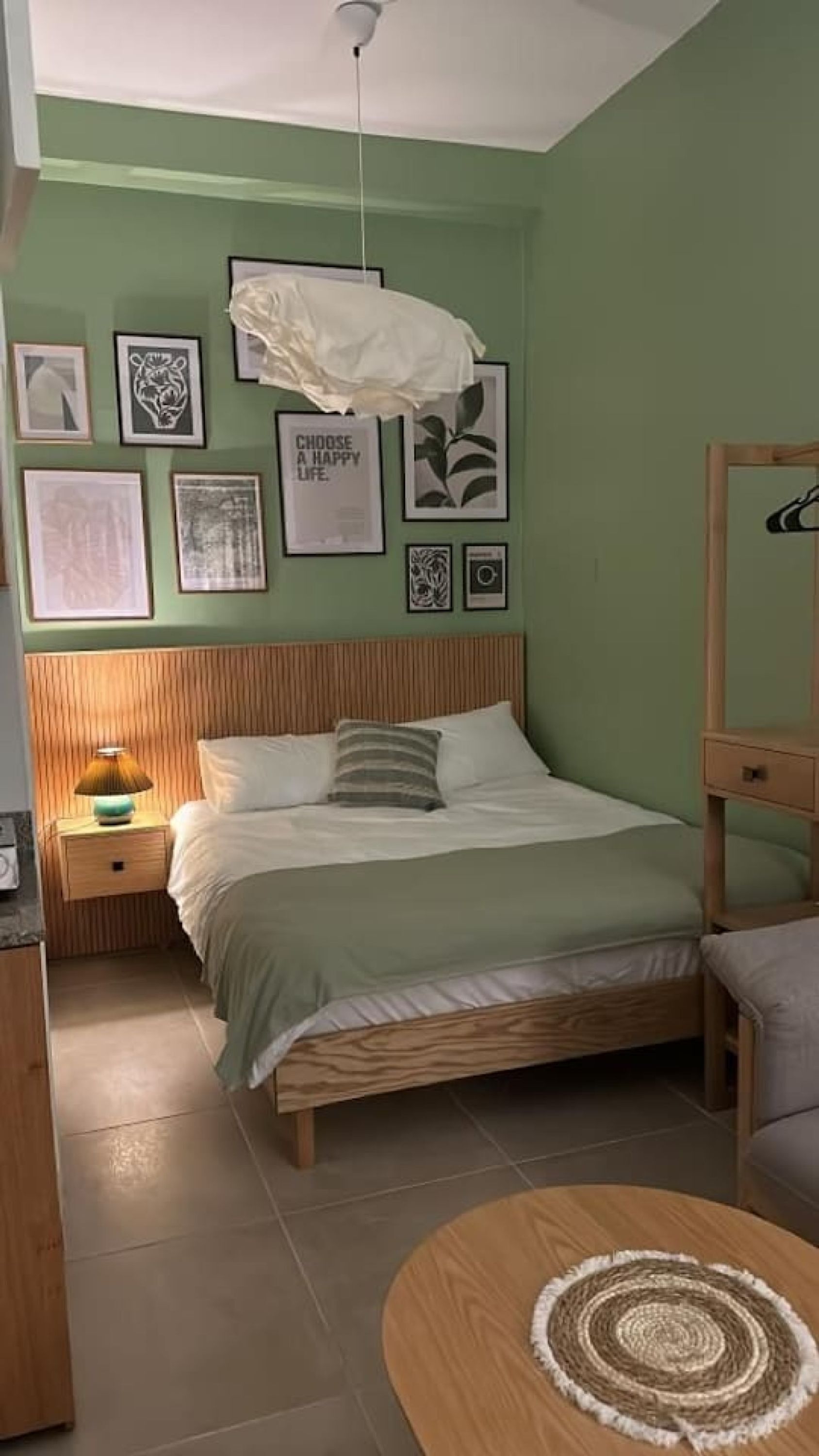 A bedroom with a bed, a bedside table with a lamp, a round wooden table, and a mirror.
