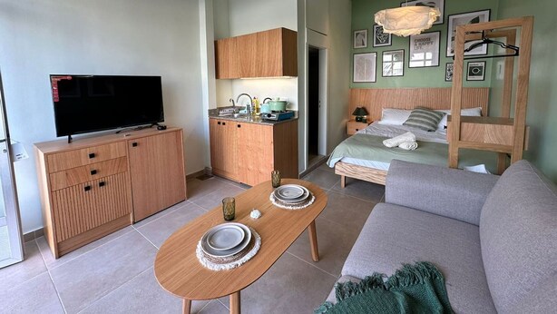 A modern hotel room with a flat-screen TV, wooden furniture, a kitchenette, a bed with a green headboard, and a hanging light fixture. - Batroun s Lovely Studio (Batroun)