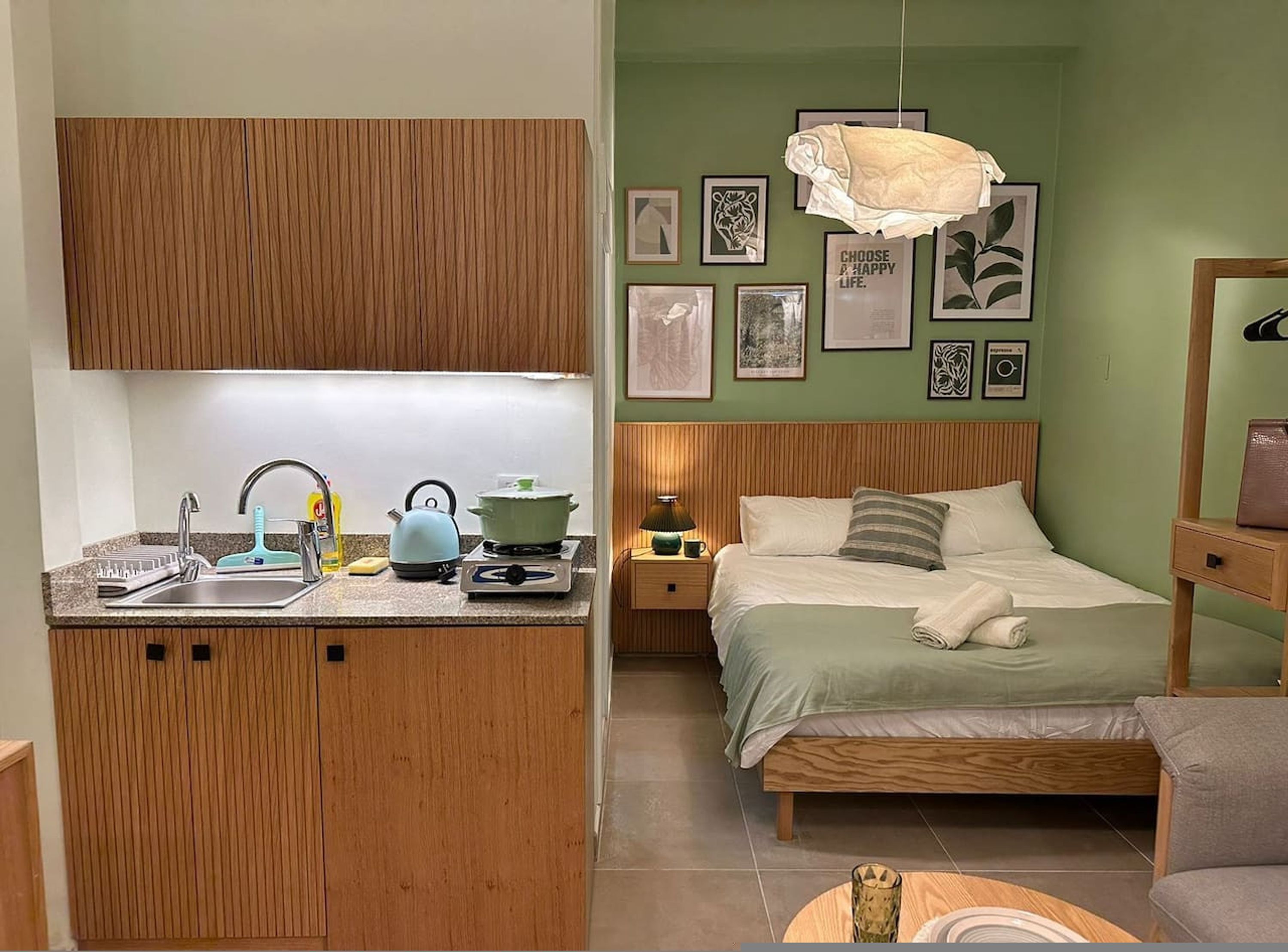 A compact bedroom with a wooden bed, a bedside table, and a nightstand. The room has a green wall with framed pictures and a hanging light fixture.