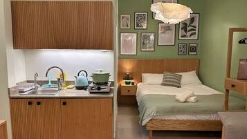 A compact bedroom with a wooden bed, a bedside table, and a nightstand. The room has a green wall with framed pictures and a hanging light fixture.