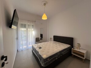 A bedroom with a bed, a TV mounted on the wall, a bedside table with a lamp, and a window with sheer curtains. - Loutraki s Seaside Serenity 2 Bdrm (Loutraki-Agioi Theodoroi)