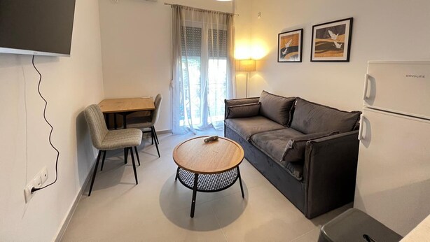 En kompakt opholdsstue med en sofa, spisebord, lille rundt bord, tv og køleskab.