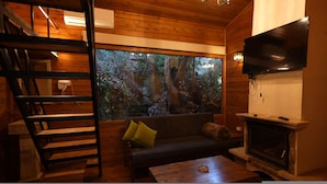 A cozy room with a wooden interior, a large window with a forest view, a flat-screen TV mounted on the wall, and a fireplace. - Keserwan s Hillside Resort and Venue (Aazra Wil Aazr)