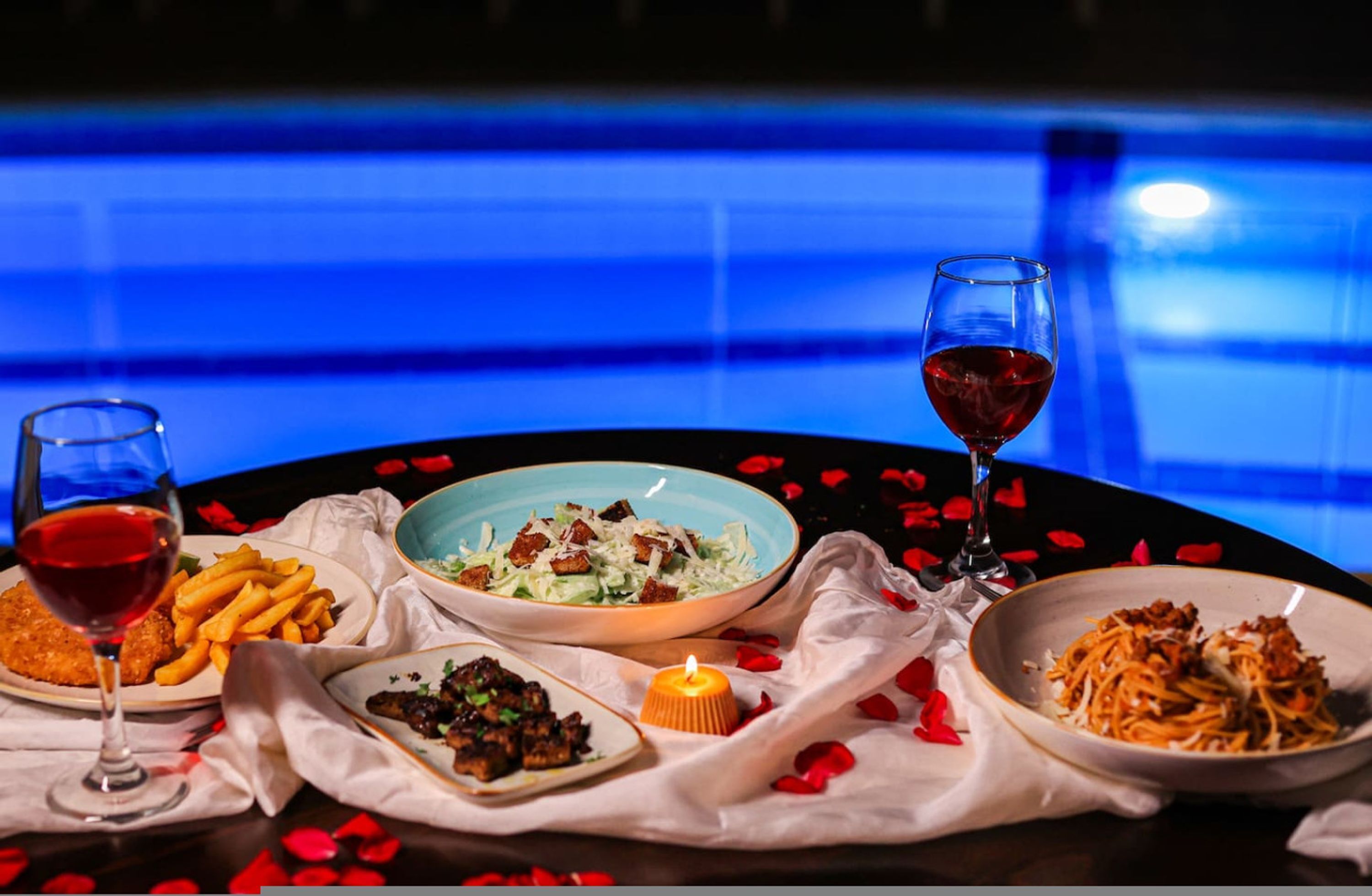 Una mesa servida con diversos platos, una vela y copas de vino tinto, con pétalos de rosa esparcidos alrededor.