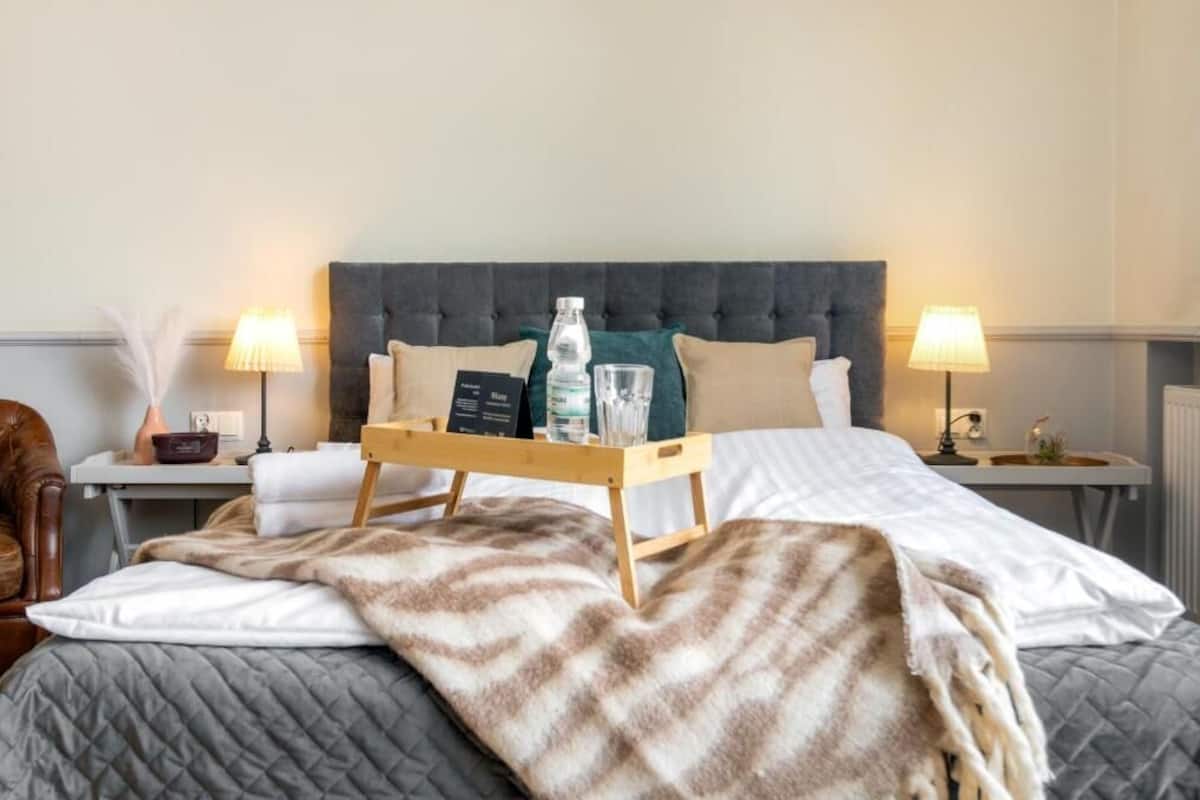 Une chambre d’hôtel avec un grand lit, une table de chevet avec une lampe, un plateau avec une bouteille et des verres, et une chaise.