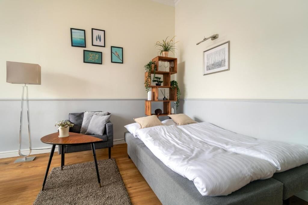 A modern bedroom with a bed, a small round table with a plant, a grey sofa, and a floor lamp. There are framed pictures on the wall and a wooden shelf with plants.
