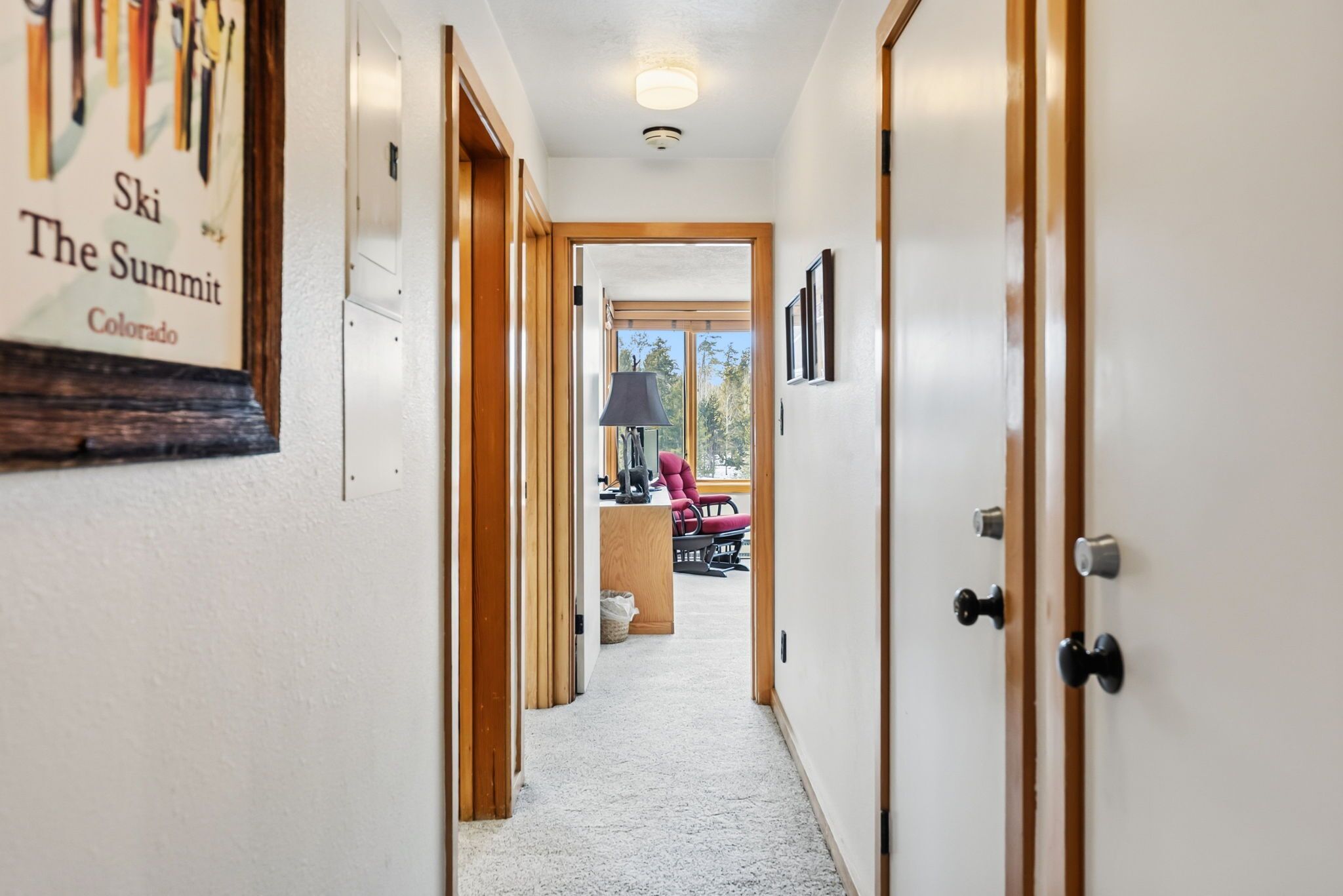 A hallway with a wooden door on the right, a framed sign on the left, and a view of a room with a chair and a lamp in the background.